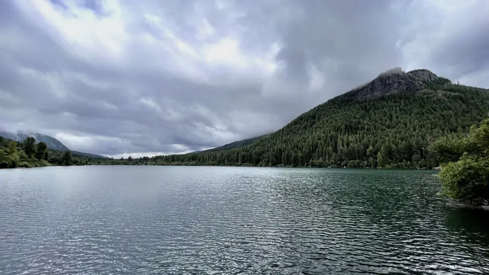 A calm lake stretches beneath a cloudy sky, bordered by dense forest and a steep rocky mountain.