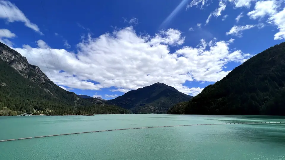 A turquoise lake stretches between forested mountains beneath a vivid blue sky filled with white clouds.