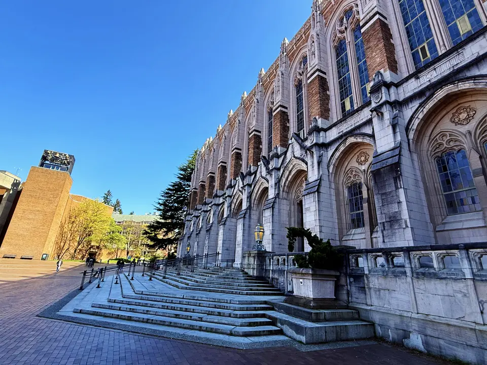 A grand Gothic-style brick and stone building with tall arched windows rises beside broad steps under a clear blue sky.