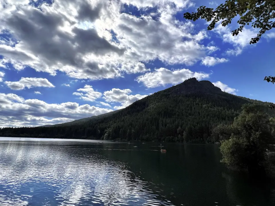 A forested mountain rises above a rippling lake beneath dramatic clouds in a bright blue sky.