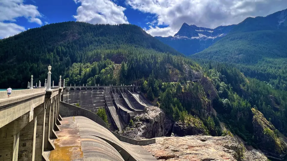 A concrete dam with a spillway stands amid forested mountains beneath a cloud-filled blue sky.