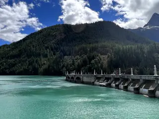 A concrete dam spans turquoise water beneath a forested mountain and a cloud-filled blue sky.