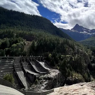 A concrete dam spillway sits beneath forested mountain slopes and a snow-streaked peak under dramatic clouds.