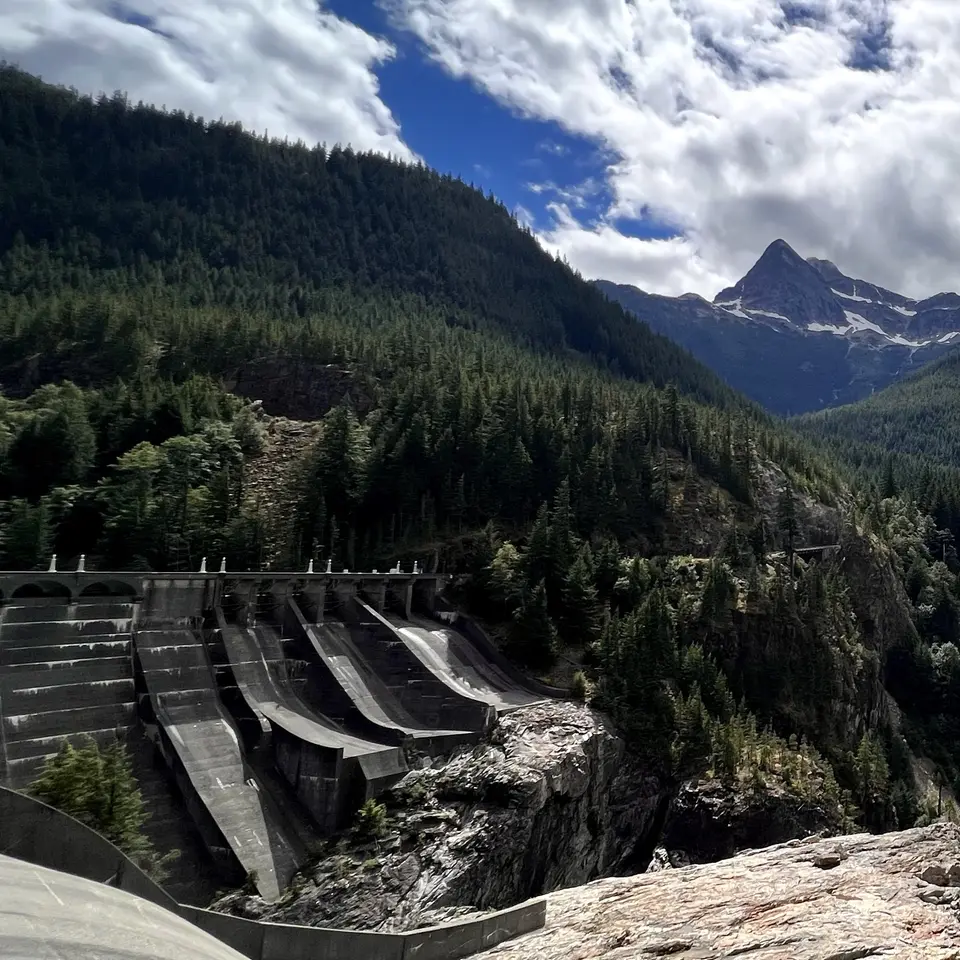 A concrete dam spillway sits beneath forested mountain slopes and a snow-streaked peak under dramatic clouds.