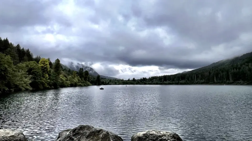 A rippling lake stretches between forested hills beneath a low blanket of gray clouds, with small boats on the water.