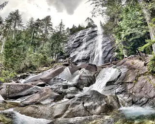 A waterfall cascades over broad dark rocks through a dense evergreen forest, with several hikers standing near the falls.