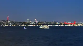 The illuminated 15 July Martyrs Bridge and Istanbul skyline stretch across dark water at night, with boats passing in the foreground.
