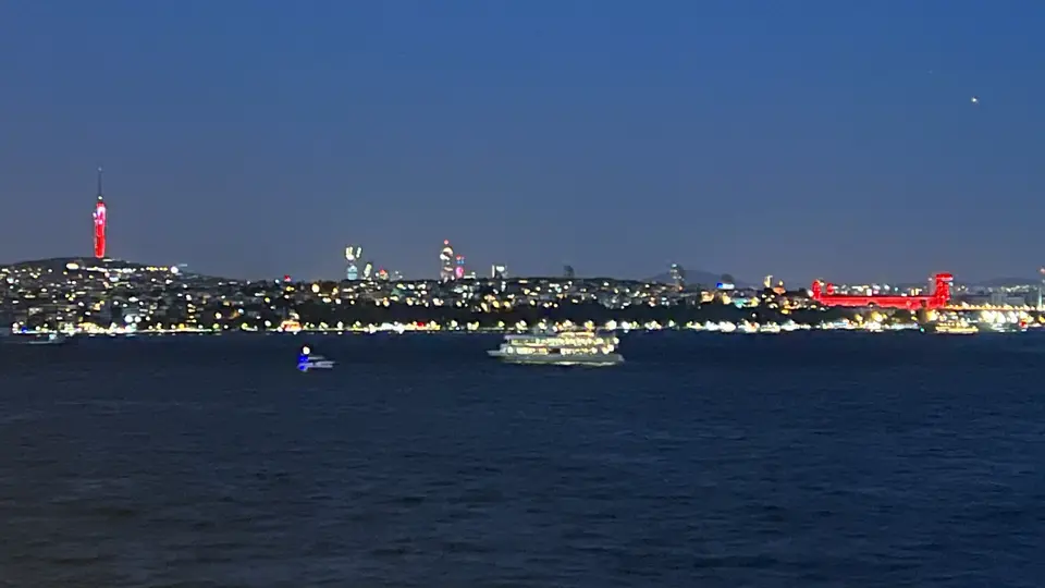 The illuminated 15 July Martyrs Bridge and Istanbul skyline stretch across dark water at night, with boats passing in the foreground.
