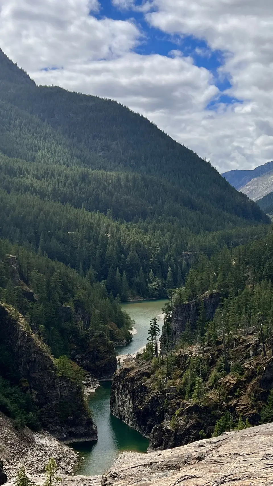 A turquoise river winds through steep rocky cliffs beneath a densely forested mountain and scattered clouds.