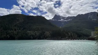Turquoise water stretches below a forested mountain range with patches of snow beneath a cloudy sky.