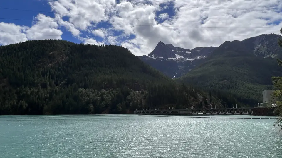 Turquoise water stretches below a forested mountain range with patches of snow beneath a cloudy sky.