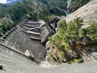 A massive curved concrete dam spans a rocky forested gorge above green water.