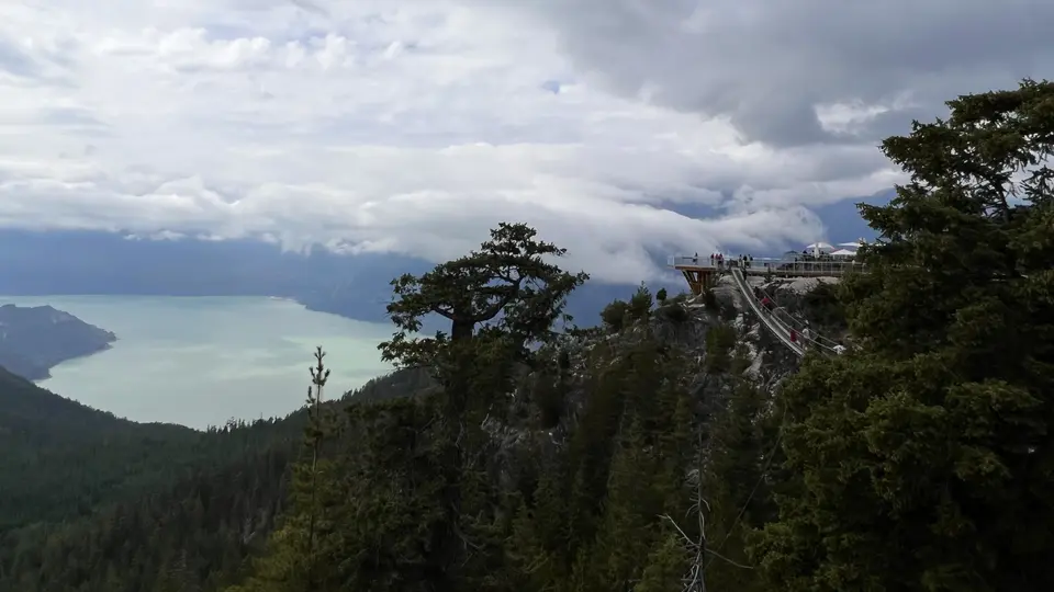 A viewing platform clings to a forested rocky mountainside overlooking a pale lake beneath heavy clouds.