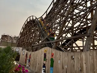 A wooden roller coaster train carrying riders climbs a steep track above a fence decorated with colorful figures.
