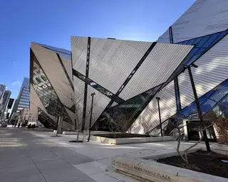 An angular modern building with metal panels and glass walls rises beside a paved plaza under a clear blue sky.