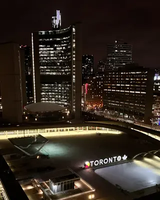 A nighttime view of illuminated downtown high-rise buildings overlooks the TORONTO sign in Nathan Phillips Square.