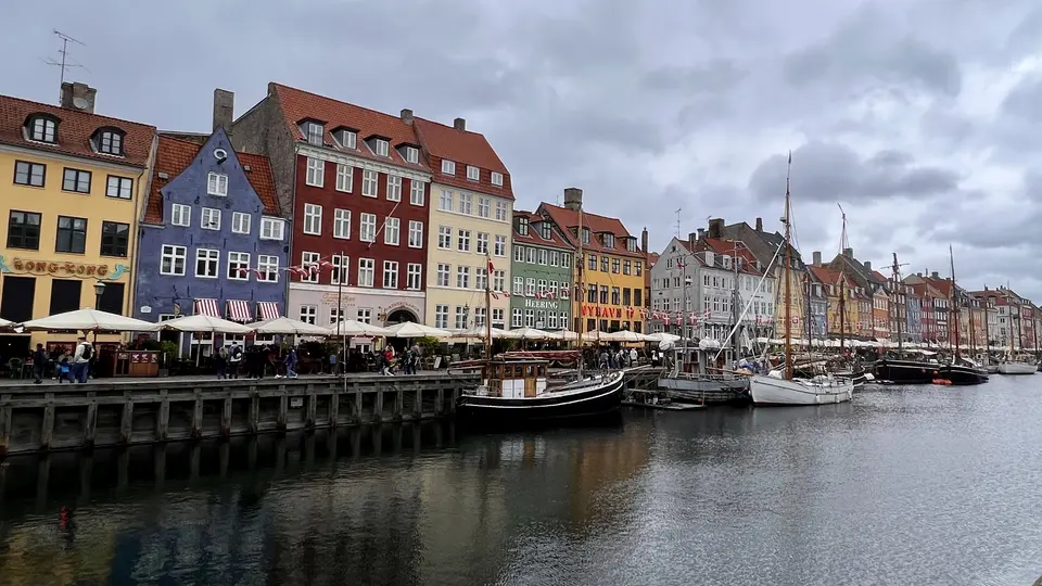 Colorful historic waterfront buildings and outdoor cafés line a harbor filled with moored boats under a cloudy sky.