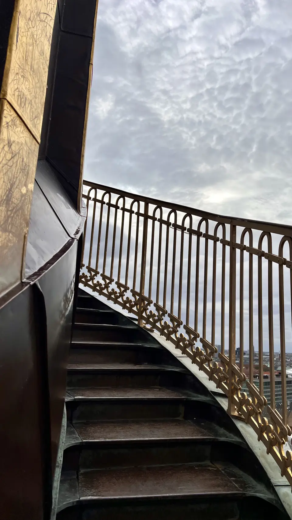 A curved outdoor staircase with an ornate metal railing rises beside a graffiti-marked wall beneath a cloudy sky.