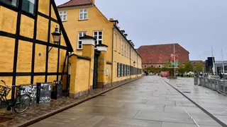 Yellow historic buildings line a wet paved walkway beside a bicycle and a waterfront railing.