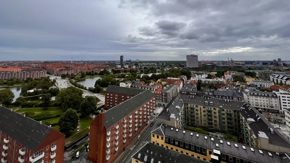 A high-angle view overlooks dense city blocks, a green park, and a small body of water beneath a cloudy sky.