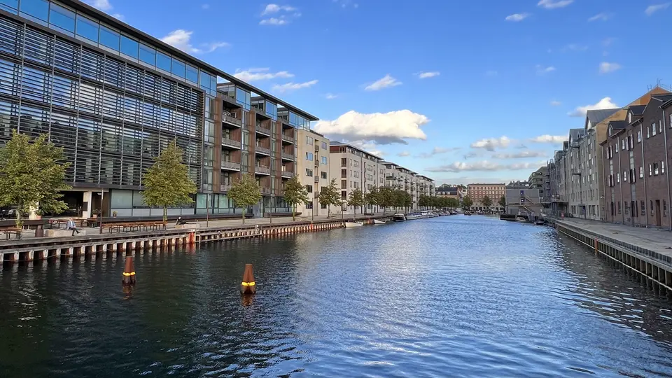 A calm canal runs between modern glass-fronted and brick apartment buildings beneath a blue sky with scattered clouds.