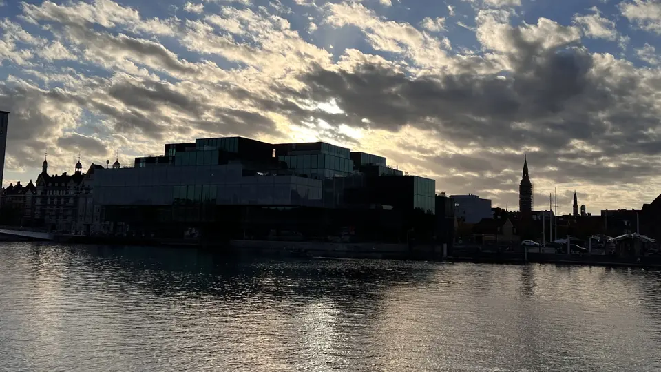 A silhouetted waterfront skyline with modern and historic buildings stands beneath a dramatic cloud-filled sunset sky.