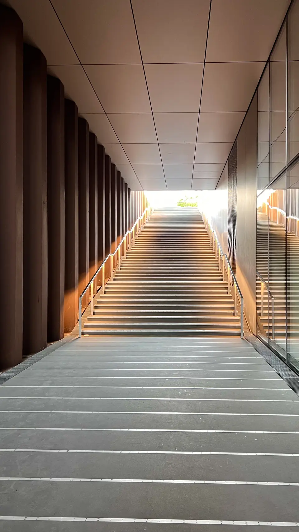A broad modern staircase rises through a covered passage lined with dark vertical columns and reflective glass walls.