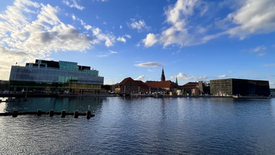 Modern waterfront buildings and a church steeple stand across rippling water beneath a blue sky with scattered clouds.