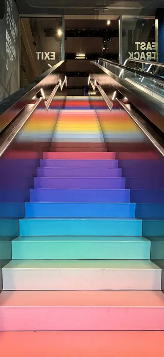 A broad staircase illuminated in rainbow colors rises between metal handrails beside an escalator, with reversed EXIT and EAST TRACK signs visible in glass reflections.