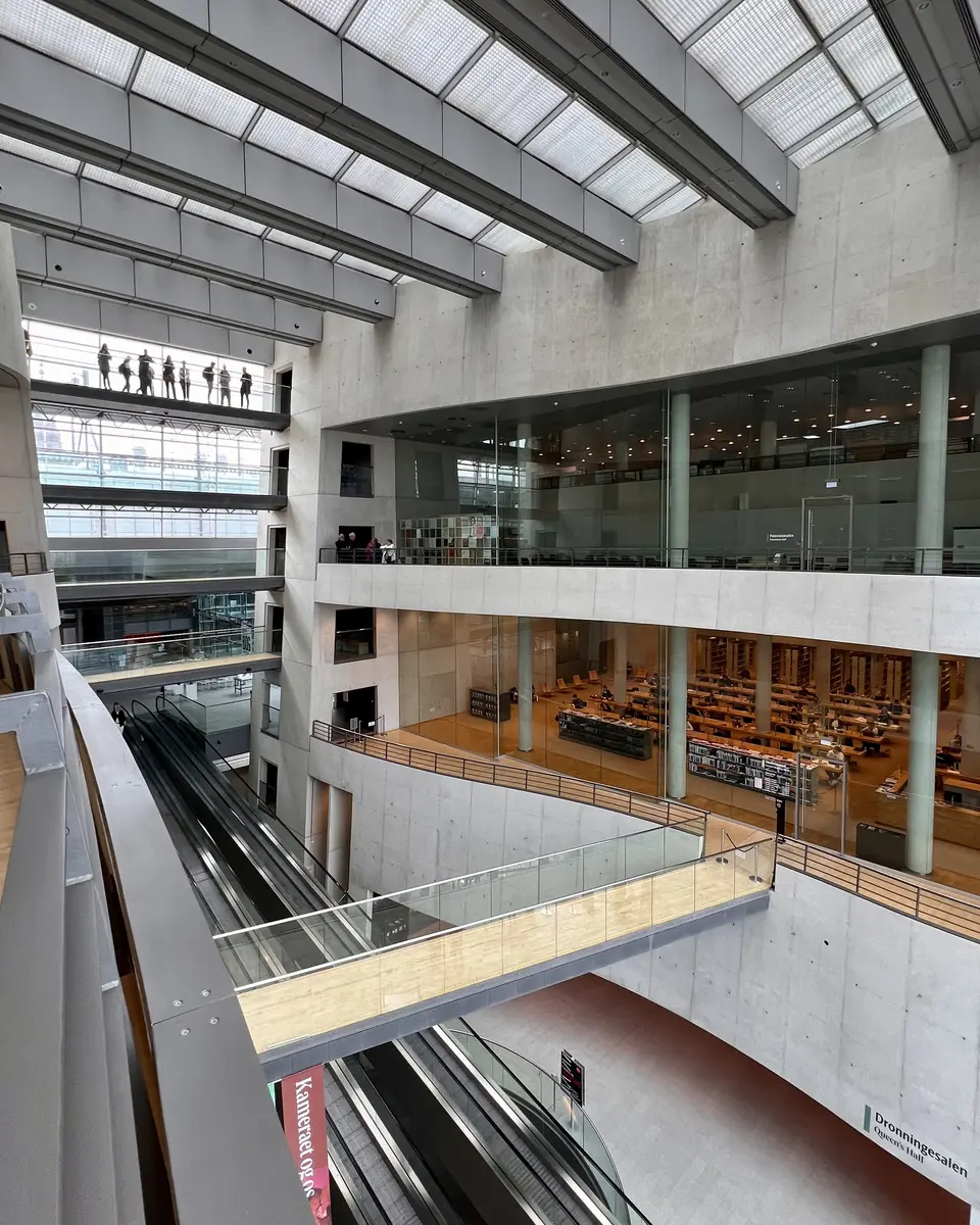 A spacious modern library atrium with glass railings, escalators, concrete walls, and multiple levels of bookshelves beneath a skylit ceiling.