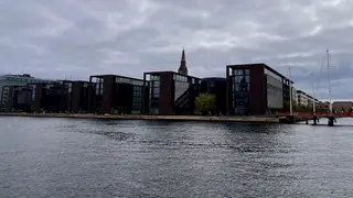 Modern dark-framed glass buildings line a waterfront beneath a cloudy sky, with a church spire rising behind them.