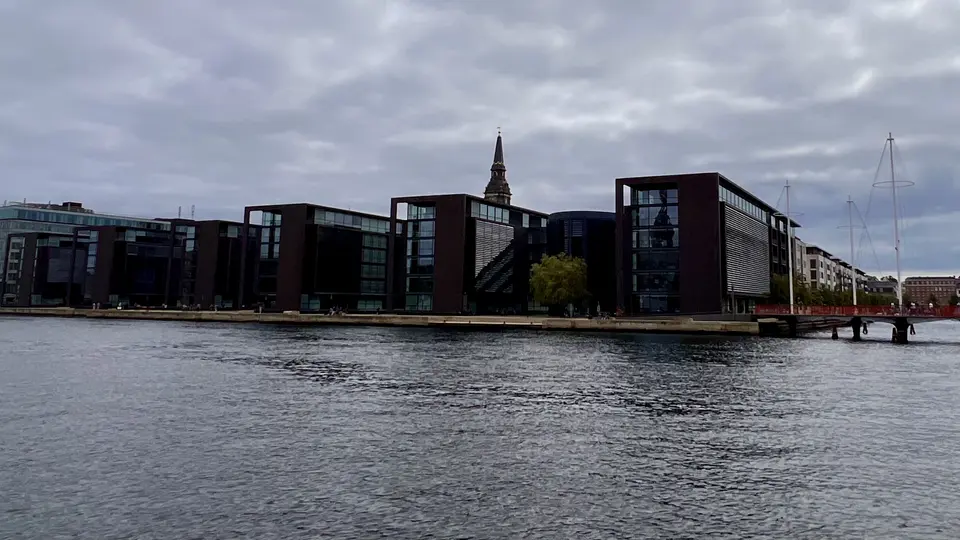 Modern dark-framed glass buildings line a waterfront beneath a cloudy sky, with a church spire rising behind them.