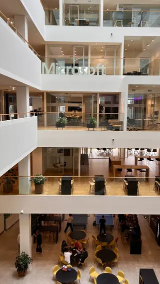 A multilevel Microsoft office atrium contains glass-walled meeting rooms, worktables, and people gathered around café tables below.