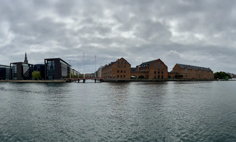 Brick waterfront buildings and modern glass offices line a canal beneath a cloudy sky.