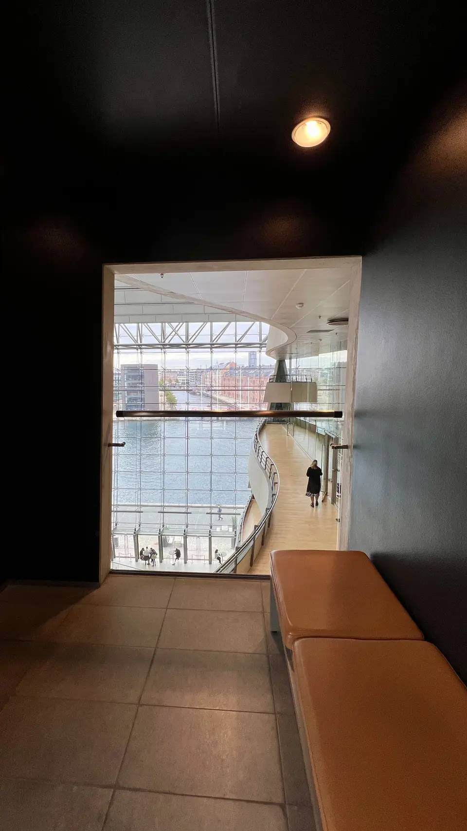 A tiled interior with an orange bench overlooks a curved glass-walled corridor beside the water.