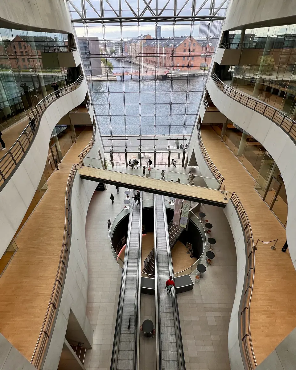 A multilevel glass-walled atrium with curved balconies and escalators overlooks a waterfront of brick buildings.