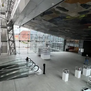 A spacious glass-walled atrium with escalators, display cases, and a dark abstract ceiling mural overlooks a city street.