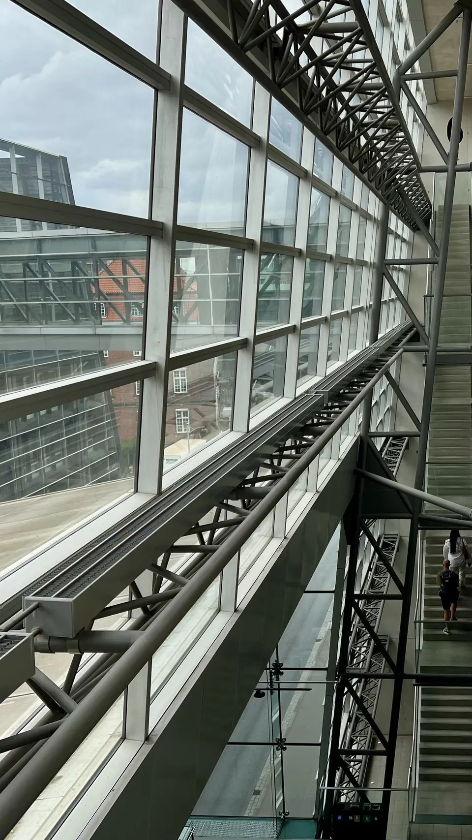 A multi-level interior atrium features large glass windows, steel trusses, and a staircase beside an elevated walkway.
