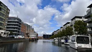 Boats are moored along a canal lined with modern apartment buildings beneath a cloudy blue sky.