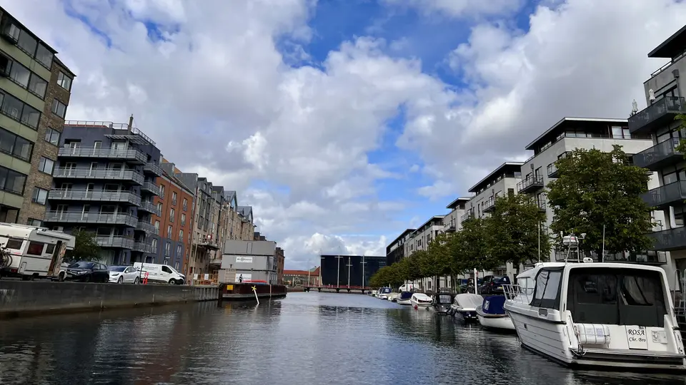Boats are moored along a canal lined with modern apartment buildings beneath a cloudy blue sky.