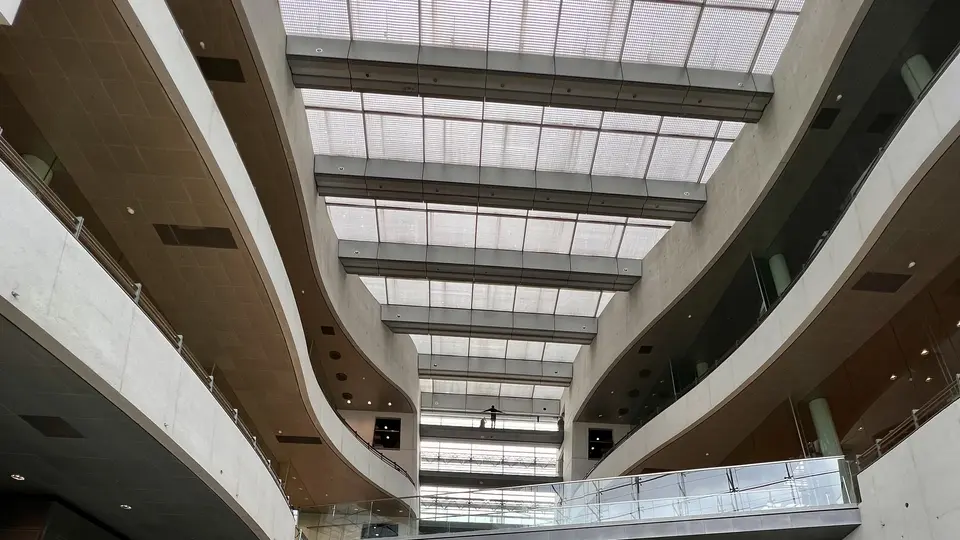 A modern multi-story interior atrium features curved balconies beneath a large gridded skylight.