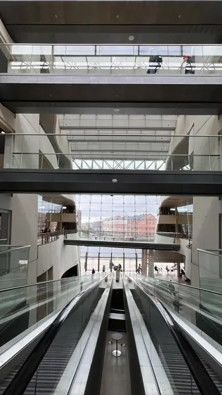 Escalators descend through a bright multilevel atrium with glass railings and large windows.