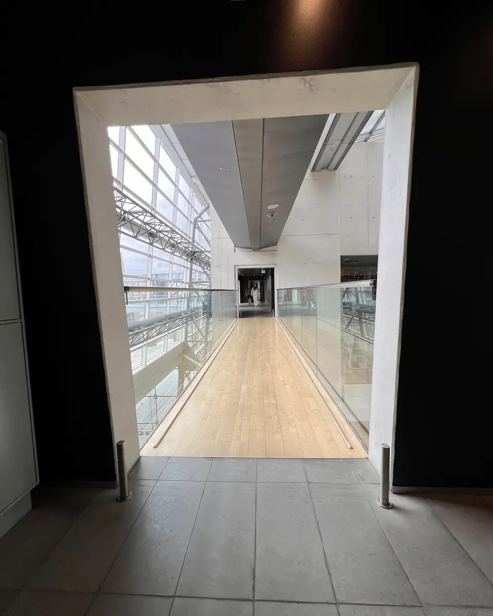 A tiled doorway opens onto a bright indoor corridor with glass railings, large windows, and a wooden floor.