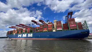 A large HMM container ship loaded with stacked shipping containers moves along the water beneath a partly cloudy sky.