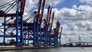 A row of towering red and blue container cranes lines a waterfront beneath a cloudy sky, with a cable-stayed bridge in the distance.