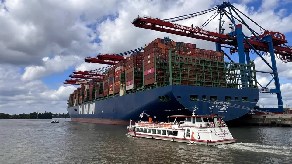 A small white passenger boat passes a container ship docked beneath cranes labeled "TERMINAL BURCHARDKAI" on a cloudy waterfront.