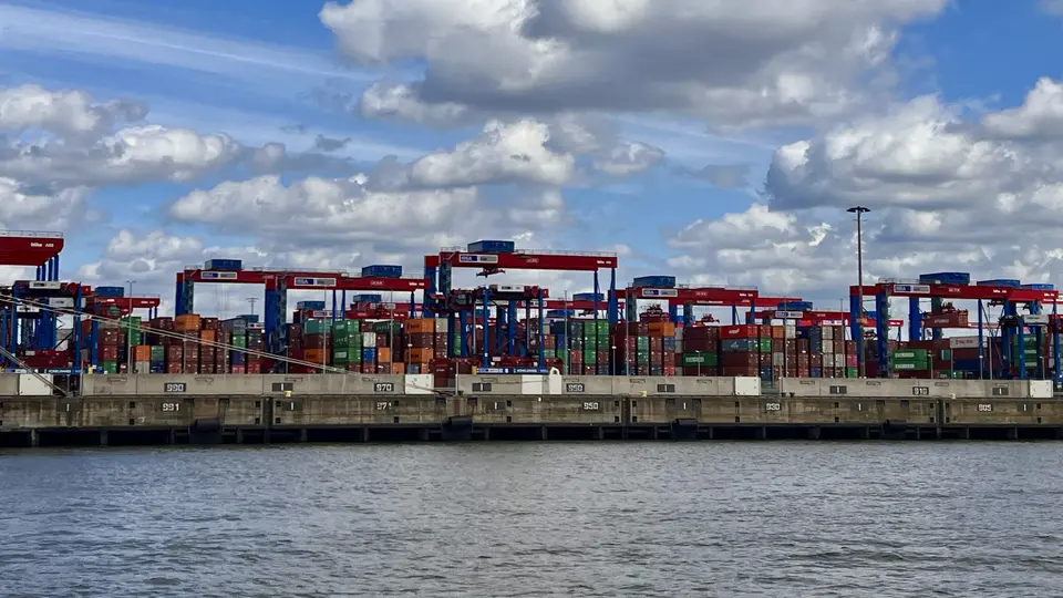 Stacked shipping containers and red loading cranes line a waterfront terminal beneath a cloudy blue sky.
