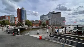 Modern high-rise buildings line a waterfront with a pedestrian bridge, docked boats, and a cloudy sky.