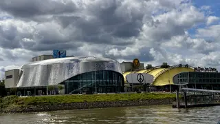 A modern waterfront complex with a silver-scaled theater facade and a yellow arched building stands beneath dramatic clouds.
