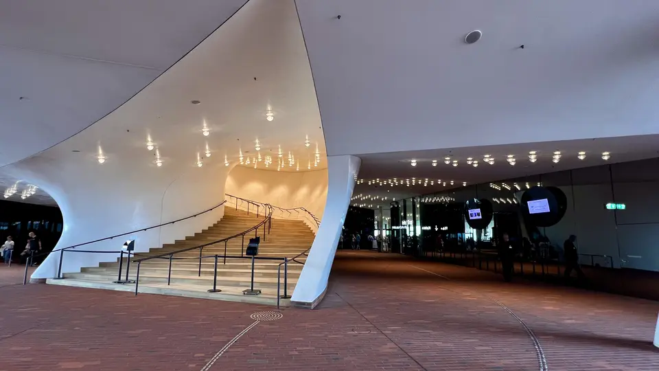 A sweeping white curved canopy frames a broad staircase with black railings leading into a brightly lit modern building entrance.